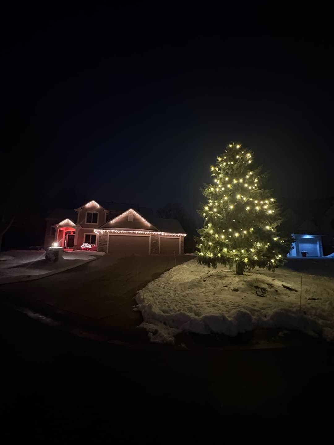 Snowy house at night with a beautifully lit Christmas tree and holiday lights.